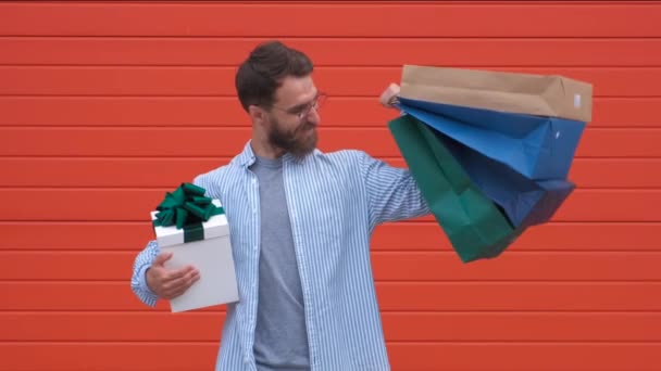 Homem com barba e bigode segura sacos de compras, fundo vermelho. Guy compras na temporada de vendas com descontos. Homem branco hipster segurando um monte de sacos de compras. Conceito de compras . — Vídeo de Stock