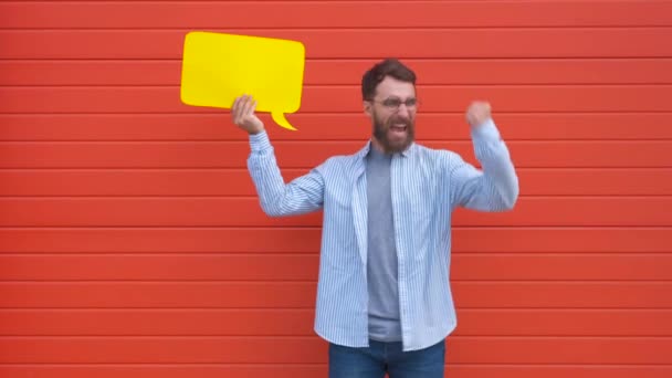Vrolijke jonge man houdt een dialoog wolk of rechthoekige gele Bubble toespraak op rode achtergrond — Stockvideo