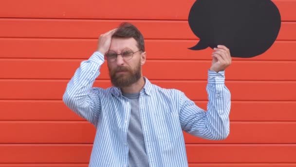 Портрет депрессивного молодого человека, стоящего и держащего черный бумажный пузырь речи над головой, на красном фоне — стоковое видео