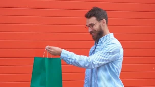 Homem com barba e bigode segura sacos de compras, fundo vermelho. Guy compras na temporada de vendas com descontos. Homem branco hipster segurando um monte de sacos de compras. Conceito de compras . — Vídeo de Stock