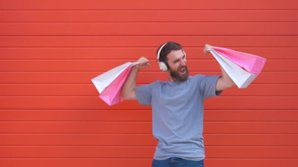 Retrato de um homem de barba hipster positivo segurando saco de compras rosa e branco no fundo vermelho. Homem ouvindo música em um fone de ouvido retro — Vídeo de Stock