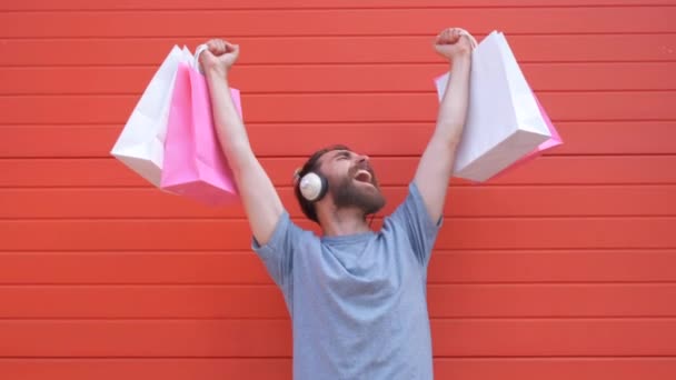 Retrato de um homem de barba hipster positivo segurando saco de compras rosa e branco no fundo vermelho. Homem ouvindo música em um fone de ouvido retro — Vídeo de Stock