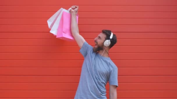 Retrato de un hombre de barba hipster positivo con bolsa de compras rosa y blanca sobre fondo rojo. Hombre escuchando música en un auricular retro — Vídeos de Stock