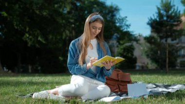 Notlarını inceleyen kız. Parkta çimlerin üzerinde oturan genç kadın, elinde açık bir defter tutuyor, uzağa bakıyor ve gülümsüyor. Mutlu öğrenci kavramı.