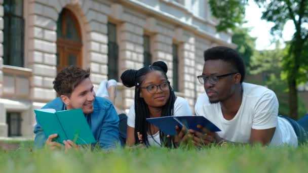 Tři studenti studující čtení poznámek spolu venku sedí na trávě — Stock video