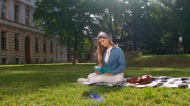 Parkta çimlerin üzerinde oturan genç kadın, elinde açık bir defter tutuyor, uzağa bakıyor ve gülümsüyor. Mutlu öğrenci kavramı.