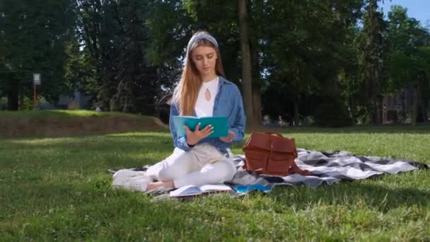 Cheerful student girl studying her notes. Young woman sitting on the grass in the park, holding an open notebook, looking away and smiling. Happy student concept. — Stock Video