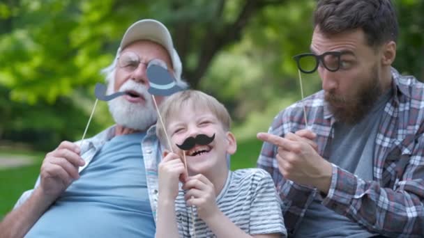 Mutlu komik aile oğlu ve baba, sahte bıyıklı dede, şapka, parkta tatil açık gözlük. İyi günler, mutluluk, dostluk, yürüyüş, tatil konsepti. — Stok video