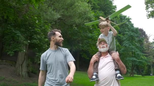 Mały chłopiec z modelem samolotu i dziadek podnoszący ręce nad zielonym parkiem na tle ciesząc się życiem i naturą. Portret szczęśliwego dziadka dając wnuka piggyback jeździć na ramionach. — Wideo stockowe
