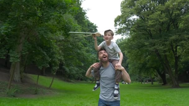 父は息子を公園に乗せて帰る。父と息子は紙飛行機を一緒に建てた。息子にピギーバックを肩に乗せた父親の肖像画。旅行休暇の夏. — ストック動画