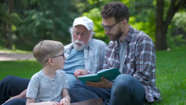 Lycklig familj av tre generationer-far, farfar och blond son sitter på gräs på Park med böcker lära sig att läsa samtidigt få redo för skolan. De är skratta, kul, spendera bra tid tillsammans — Stockvideo