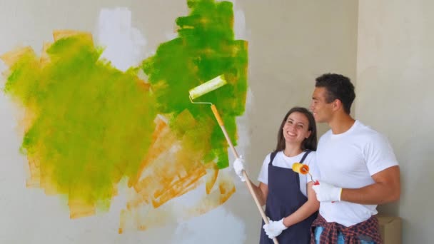 Pareja sonriente pintando su casa de verde posando juntos frente a una pared medio acabada con un rodillo y una bañera de pintura . — Vídeo de stock