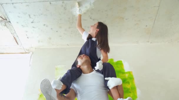 Couple is having fun redecorating their new appartment. Happy girl sits on the shoulders of the guy and paints the wall paint — Stock Video