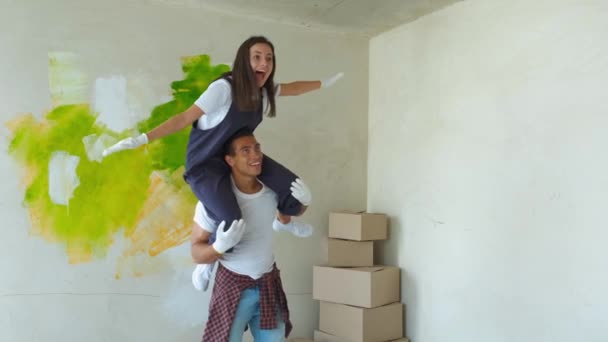 Couple is having fun redecorating their new appartment. Happy girl sits on the shoulders of the guy and paints the wall paint — Stock Video