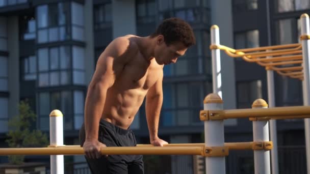 Serious strong young man in looking straight while doing pull-ups on workout area near house — Stock Video