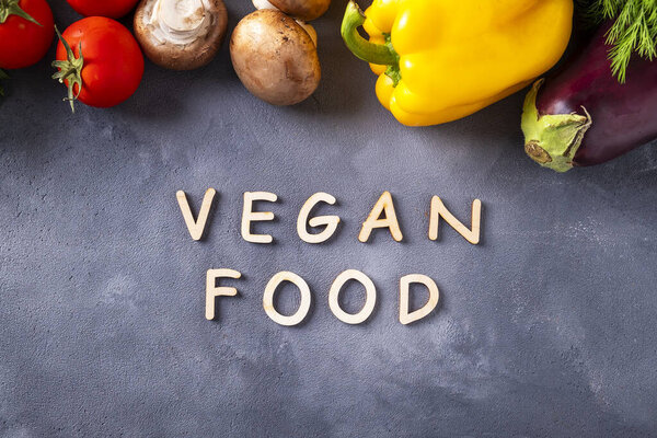 Веганская еда. Слова "Vegan food" написаны деревянными буквами на сером фоне и сырыми органическими овощами. Ингредиенты для здорового веганского питания. Вид сверху