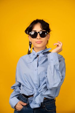 Güneş gözlüklü genç kız sarı arka planda fotoğraf için poz veriyor..