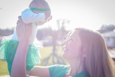closeup of a smiling mother lifting up baby in bright sunlight on St. Patrick's Day clipart
