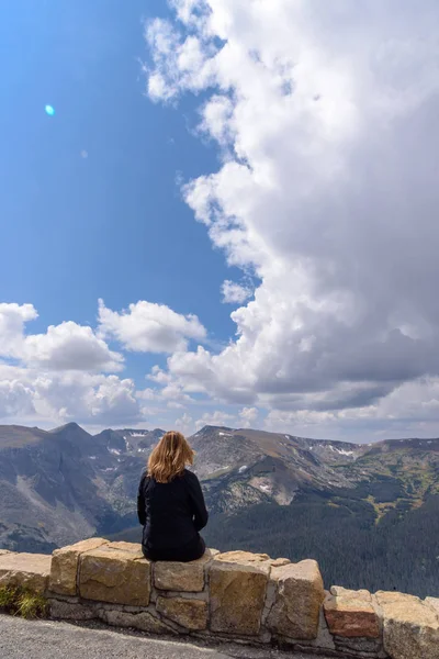 woman sitting on rock wall ledge enjoying amazing view from peak of rocky mountain range