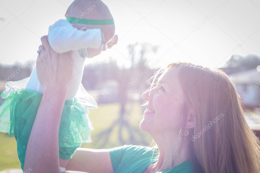 closeup of a smiling mother lifting up baby in bright sunlight on St. Patrick's Day