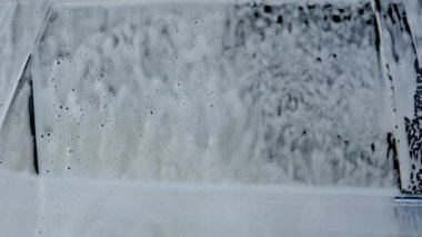 Temiz bir arabanın camından dökülen desenli beyaz sabun köpüğü. Kabarcıklar, araç yıkama işlemi sırasında soyut desenler oluşturur. Su damlaları.