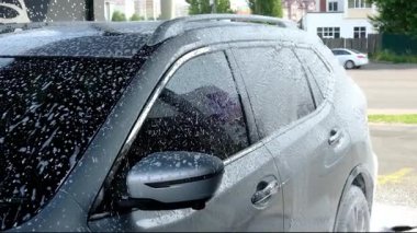 Kalın beyaz sabun köpüğü ve su kabarcıklarıyla kaplı gri bir araç. Güçlü bir basınç spreyiyle dışarıda araba temizliği..