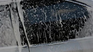 Kalın beyaz sabun köpüğü koyu renk bir araba penceresini ve yan panelini kaplar. Taze köpükler, iyice yıkadıktan sonra temiz otomotiv yüzeyine damlıyor..