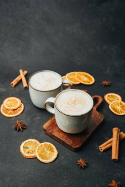 İki kupa sıcak köpüklü içecek, tarçın çubukları ve koyu renkli portakal dilimleri..