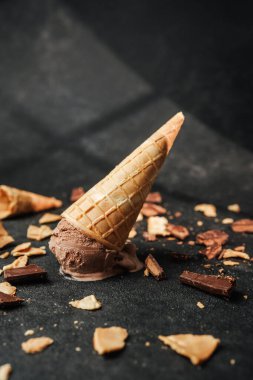 Çikolatalı dondurma külahı, üzerinde waffle kırıntıları ve çikolata parçacıkları olan karanlık bir yüzeyde yatıyor.