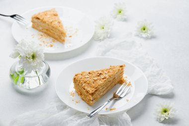 Beyaz tabakta kremalı Napolyon keki, narin çiçekler ve kumaş.