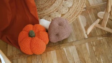 Tahta zeminde kahverengi yastığın yanında turuncu kabak peluş oyuncak, yeşil halı..