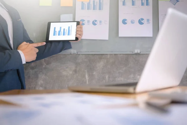 Young businessman in the suit explaining data chart on digital tablet and documents on the wall in a office.