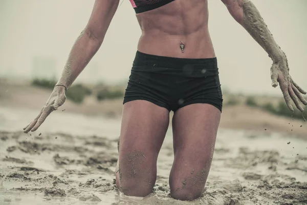 Closeup of a strong athletic woman on her knees showing six pack abs  in wet muddy puddle with mud on her hands in an extreme competitive sport