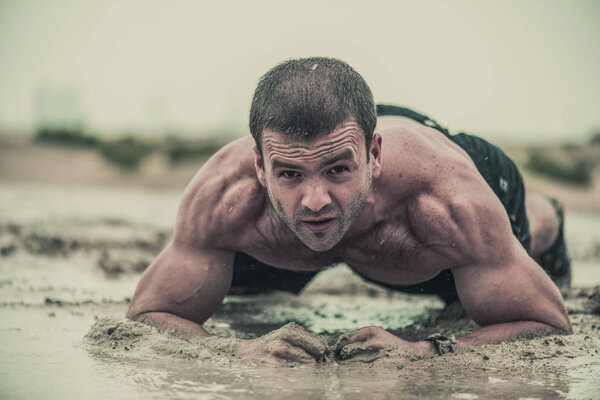 Closeup of strong, muscular, athletic man crawling in a wet, dirty muddy puddle in the rain in an extreme competitive sport 