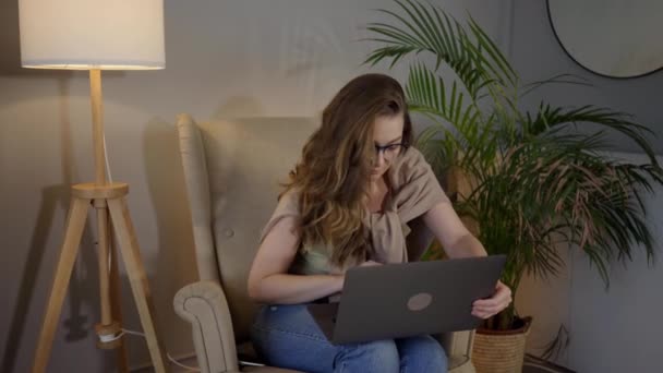 Młoda kobieta robi badania dla swojej firmy. Uśmiechnięta kobieta siedząca na sofie relaksująca się przeglądając stronę internetową zakupów online. Szczęśliwa dziewczyna przeglądająca internet w czasie wolnym w domu. — Wideo stockowe