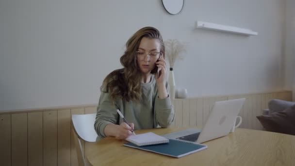 遠隔操作で働く電話で顔のない女性のテキストメッセージ。ノートパソコンとノートパッドで働く眼鏡の若い学生 — ストック動画