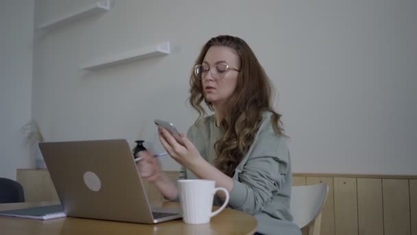 Message texte féminin sans visage sur le téléphone travaillant à distance. Jeune étudiant en lunettes de travail avec ordinateur portable et bloc-notes — Video