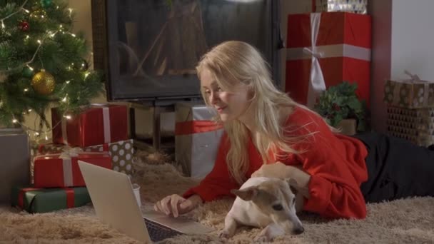 Chica feliz en Santa Sombrero de compras en línea en el ordenador portátil y sentado con un lindo perro en el hermoso árbol de Navidad de oro con luces y regalos en la habitación festiva. venta de Navidad y concepto de descuento — Vídeos de Stock