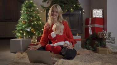 Anne ve kızı bir Noel ağacının yanında oturmuş dizüstü bilgisayar ve telefonda çizgi film izliyor ve eğleniyorlar..