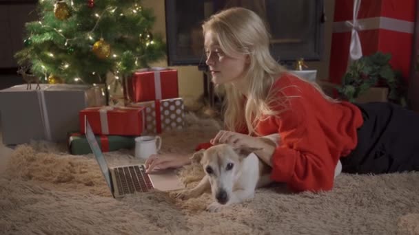 Menina feliz em santa chapéu de compras on-line no laptop e sentado com o cão bonito na árvore de Natal bonita dourada com luzes e presentes na sala festiva. venda de Natal e conceito de desconto — Vídeo de Stock