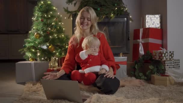 Mor och dotter sitter på golvet bredvid en julgran, tittar på tecknade serier på en bärbar dator och telefon, har kul. — Stockvideo