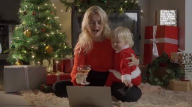 Anne ve kızı bir Noel ağacının yanında oturmuş dizüstü bilgisayar ve telefonda çizgi film izliyor ve eğleniyorlar..
