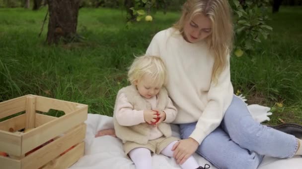 Mamma och dotter skördar äpplen och vilar på gräset. Baby med ung mamma äter äpple i höstparken. — Stockvideo