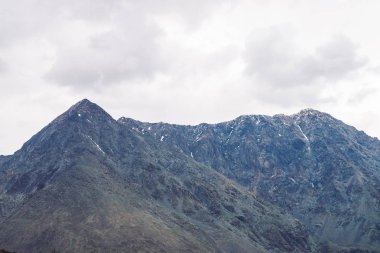 Bulutlu gökyüzünün altında kar ile dev sırt. Atmosferik sisli kayalık dağ yamacı. Bulutlu havalarda inanılmaz bir dağ sırası. Alışılmadık sisli kayalar. Yaylaların görkemli doğasının harika manzarası.