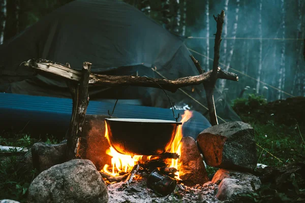 Food in cauldron is heats up on bonfire, surrounded by stones near tent. Cooking in open air in forest. Active outdoor recreation. Camping in wild at evening. Atmospheric bonfire at leisure close up.