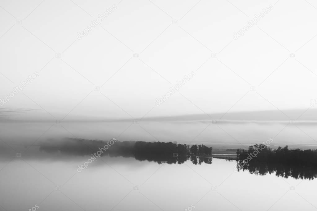 Broad mystical river flows along diagonal shore with silhouette of trees and thick fog in grayscale. Morning milky atmosphere. Minimalistic monochrome landscape of majestic nature.