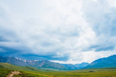 Uzak dev dağların muhteşem manzarası. Yaylalarda vadiden geçen patika. Yürüyüş yolu. Bulutlu gökyüzü altında Harika büyük dağ aralığı. Görkemli doğanın inanılmaz dramatik yeşil manzara.