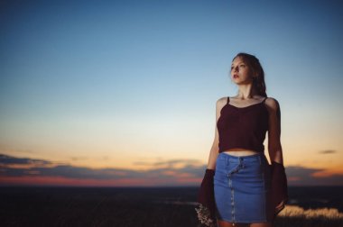 Kızıl saçlı kız güzel bir günbatımı gökyüzünün arka planında bir uçurumun eşiğinde duruyor. Grunge tarzı giysi. Elinde çiçekler.