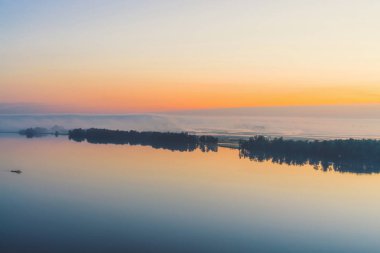 Geniş mistik nehir ağaçlar ve kalın sis silueti ile diyagonal kıyı boyunca akar. Şafak tan önce gökyüzünde altın parlıyor. Sıcak tonlarda görkemli doğanın sakin sabah atmosferik manzara.