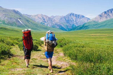 Büyük sırt çantaları ile Gezginler kar ile harika dev dağlara yeşil vadide patika üzerinde gidin. Dağlık arazide yürüyüş. Görkemli doğanın inanılmaz canlı dağ manzara. İnsanlar yükseklere tırmanıyor..